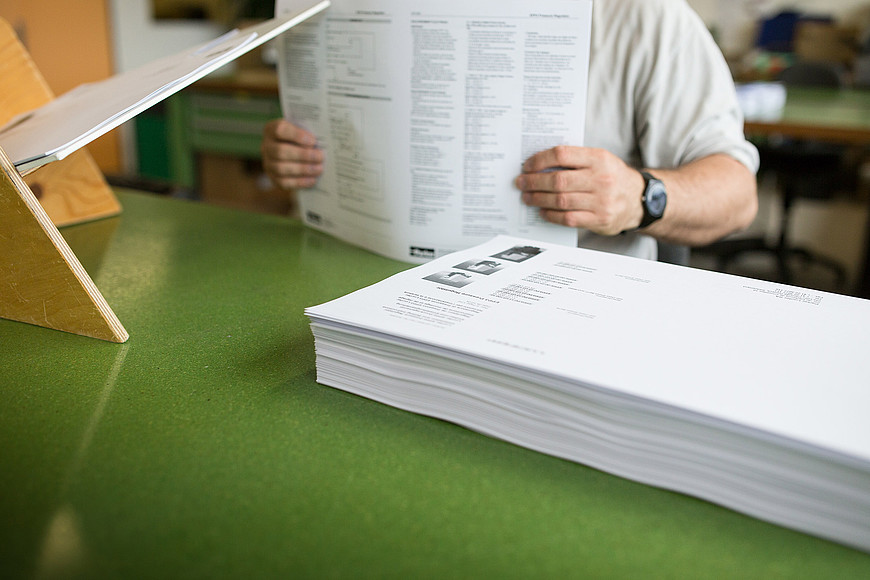 Au premier plan, une pile de papier imprimé, au second plan une personne debout tient dans ses mains des feuilles de papiers.