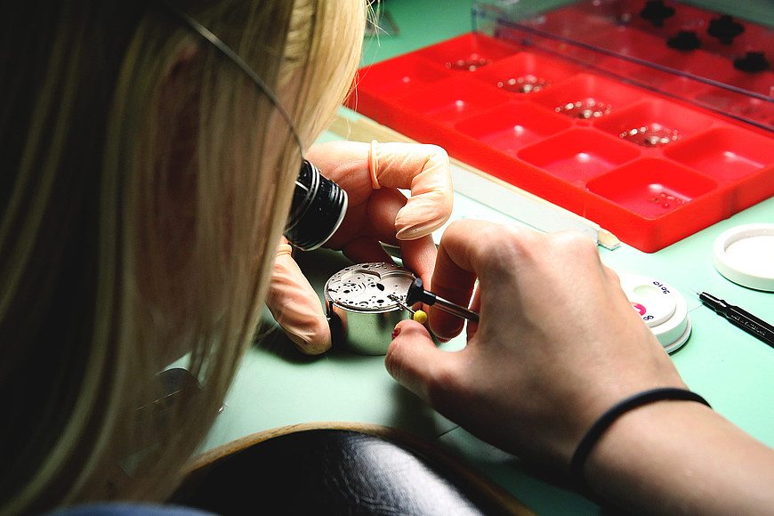 Une jeune femme blonde est concentrée et travaille sur un boîtier de montre, elle porte une loupe œil d'horloger